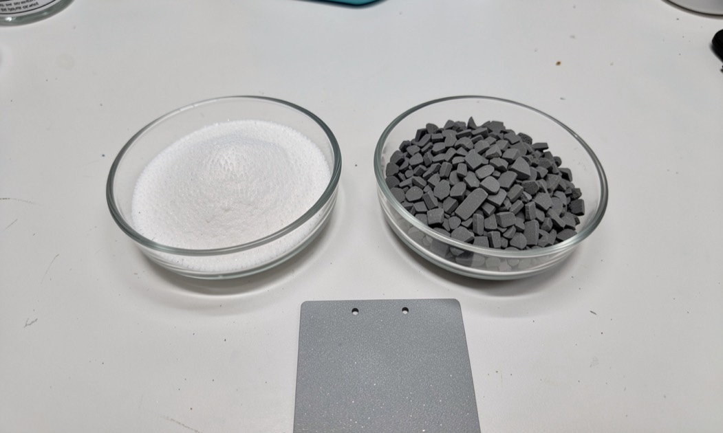White polymer powder and dark ceramic grit in two laboratory dishes beside a coated test panel